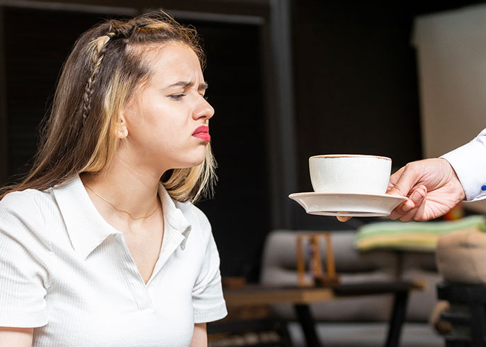 Mujer con expresión de desagrado rechazando una taza de café servida por una persona, mostrando una reacción poco inteligente.