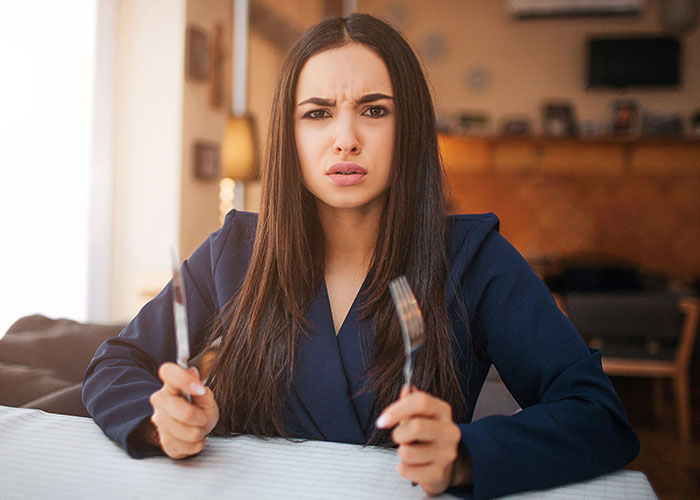 Mujer confundida sosteniendo tenedor y cuchillo en mesa, mostrando una expresión que refleja lo difícil que es creer su poca inteligencia.