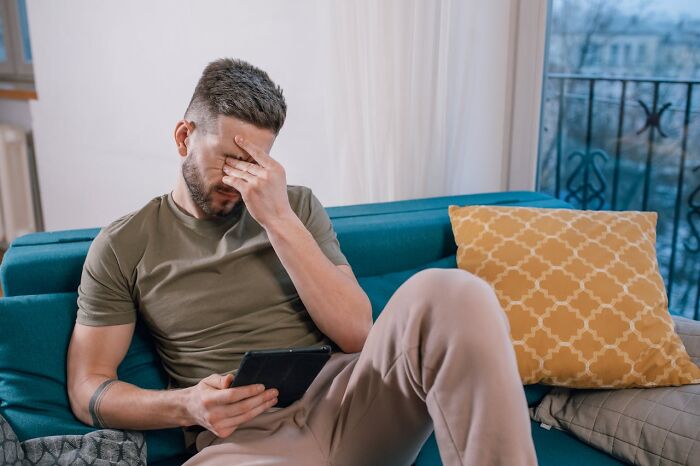 Hombre sentado en sofá con expresión de frustración, sosteniendo una tablet, simbolizando mundo que se desmorona accidentalmente.