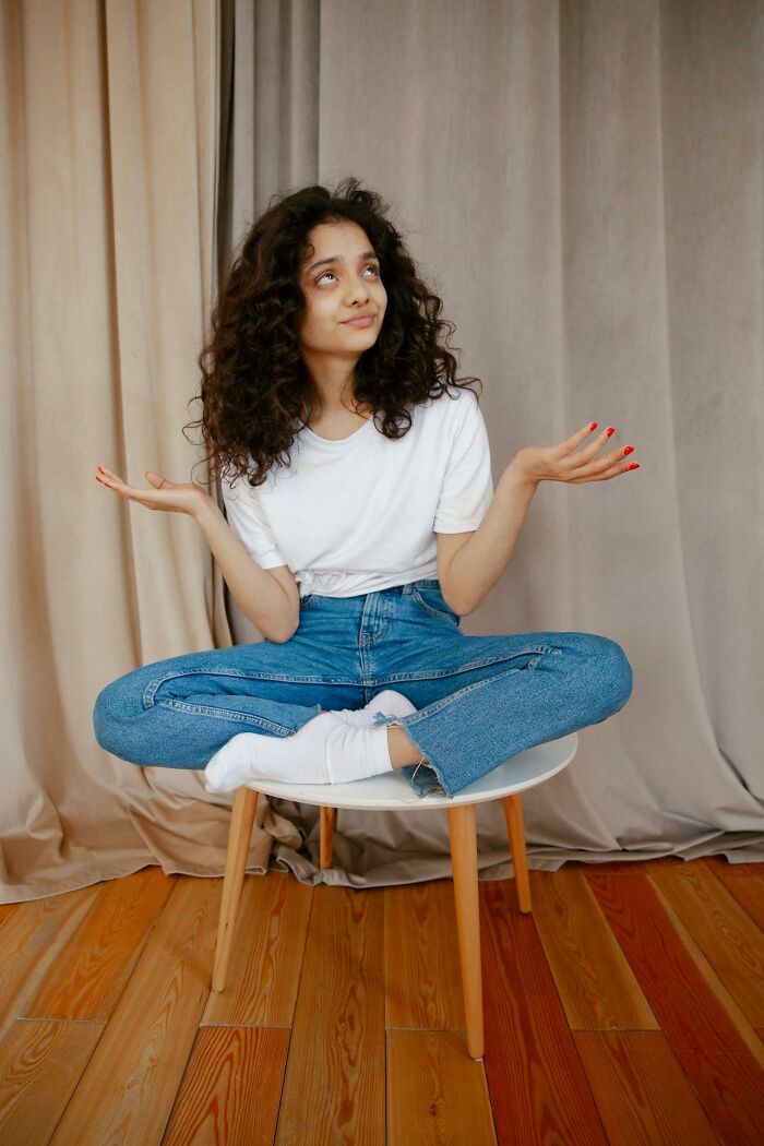 Mujer con cabello rizado sentada con piernas cruzadas, expresando confusión en un ambiente interior minimalista, reflejando impacto accidental.