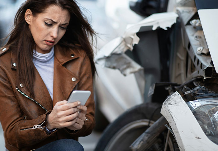 Mujer angustiada revisando su teléfono junto a un auto accidentado, reflejando culpa y arrepentimiento por arruinar vidas.
