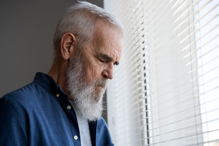 Hombre mayor con barba mirando por la ventana, reflexionando sobre las consecuencias de mentiras para otras personas.