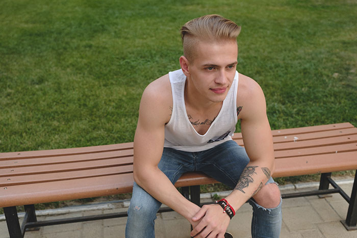 Joven sentado en banco al aire libre, con tatuajes, mostrando consecuencias de mentiras en otras personas.