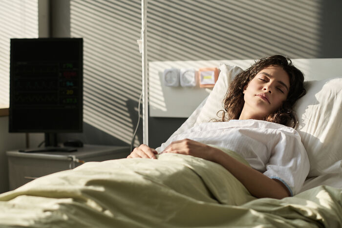 Paciente con expresión de malestar en cama de hospital junto a monitor médico en sala iluminada por luz natural.