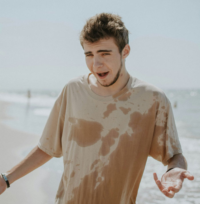 Joven en la playa con camiseta manchada, mostrando expresión confundida en momentos hilarantes de desconocimiento común.