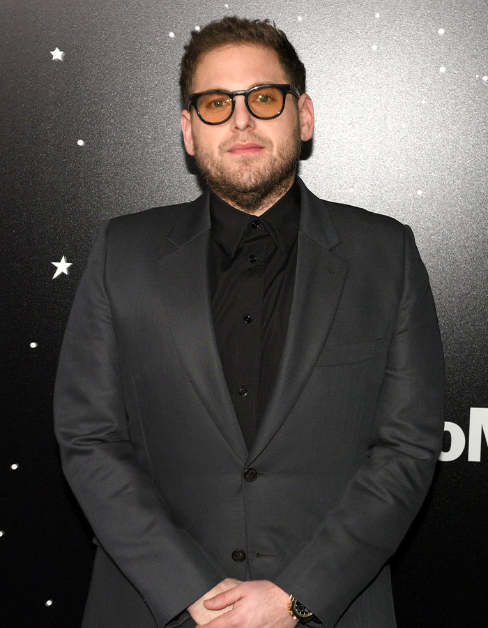 Actor masculino con gafas y traje gris, uno de los famosos cuya estatura real sorprende a todos.