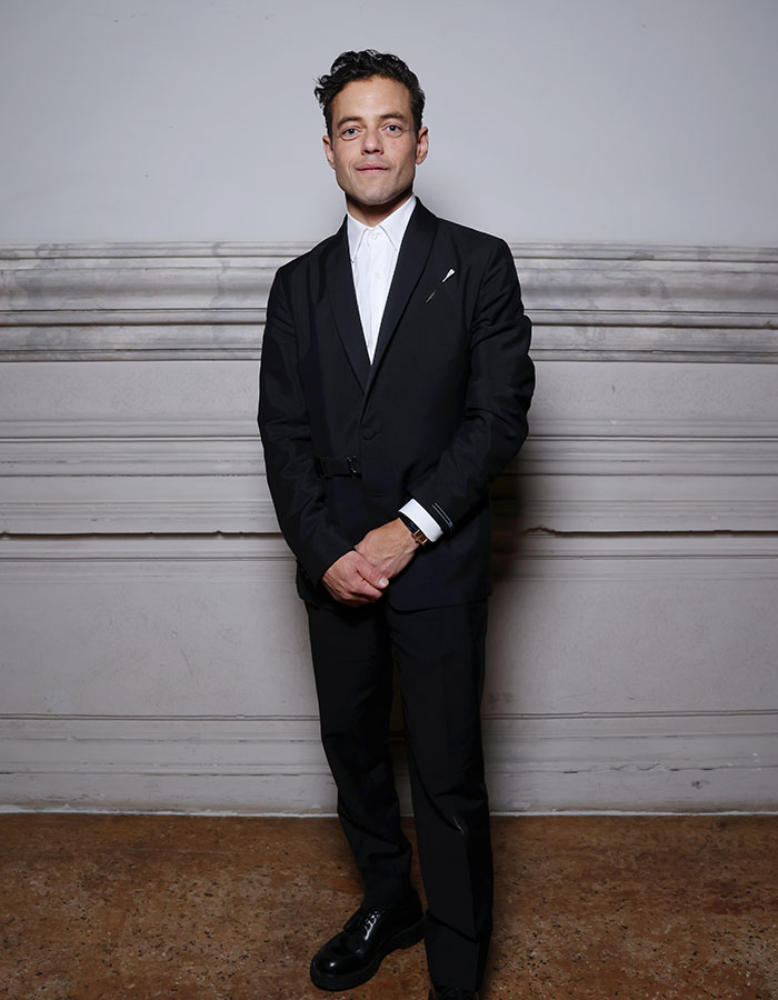 Hombre joven vestido de traje negro posando frente a pared blanca, ejemplo de estrellas masculinas y su altura real sorprendente.