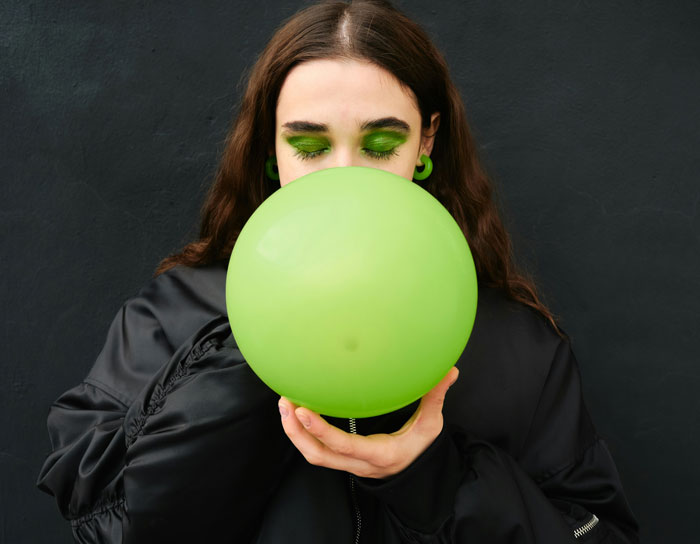 Mujer con sombra y pendientes verdes inflando un globo verde, imagen divertida sobre desconocimiento de conocimientos comunes.