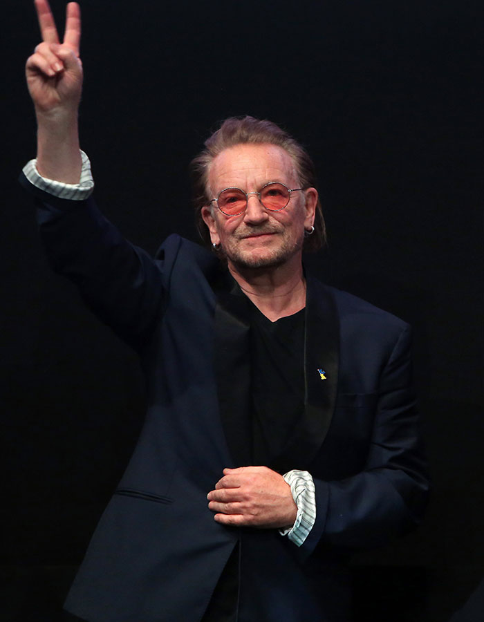 Hombre famoso con gafas rosas y chaqueta azul saludando, ejemplo de estrellas masculinas y su altura real sorprendente.
