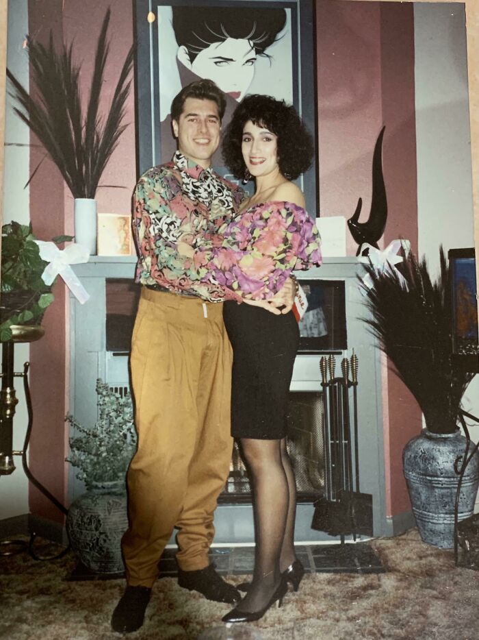 Pareja con ropa colorida de moda ochentera posando frente a chimenea, captura que muestra estilo salvaje de los 1980s.