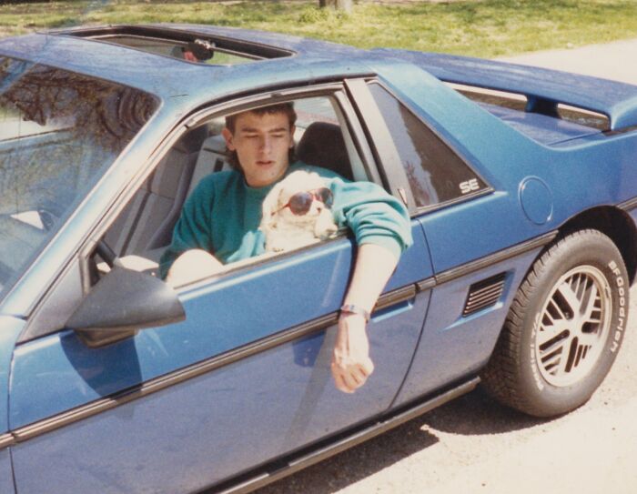 Joven de los años 1980 conduciendo un auto deportivo azul con un perro usando gafas de sol en el asiento delantero.
