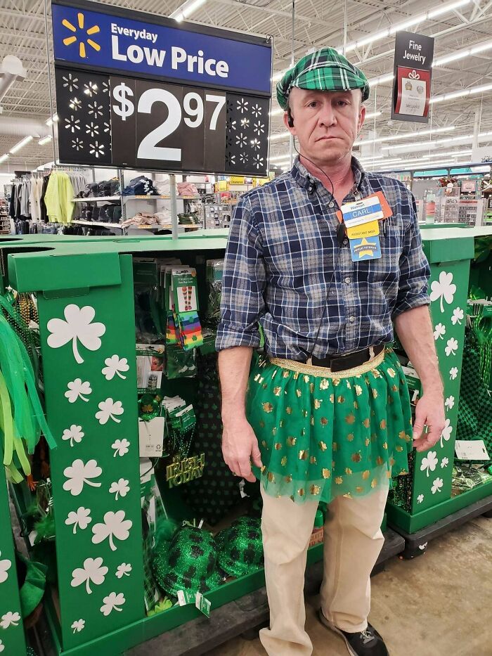 Empleado de Walmart vestido con falda verde y gorra a cuadros, posando junto a artículos de decoración de tréboles.