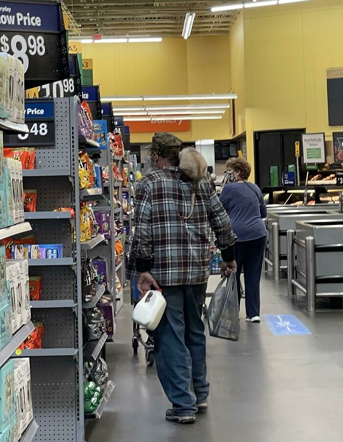 Hombre con camisa a cuadros en Walmart llevando un envase de leche y una bolsa mientras compra en pasillo de snacks.