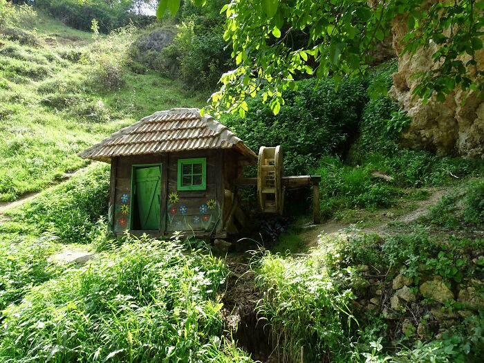 Pequeña casa cottagecore con rueda de agua rodeada de vegetación en paisaje rural tranquilo ideal para sueños cottagecore.