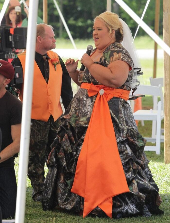 Novia con vestido de boda único en camuflaje y lazo naranja en una ceremonia al aire libre.