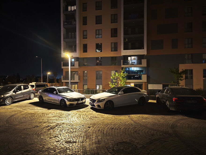 Coches estacionados ocupando espacios en edificio de apartamentos, dificultando encontrar lugar de parking en la noche.