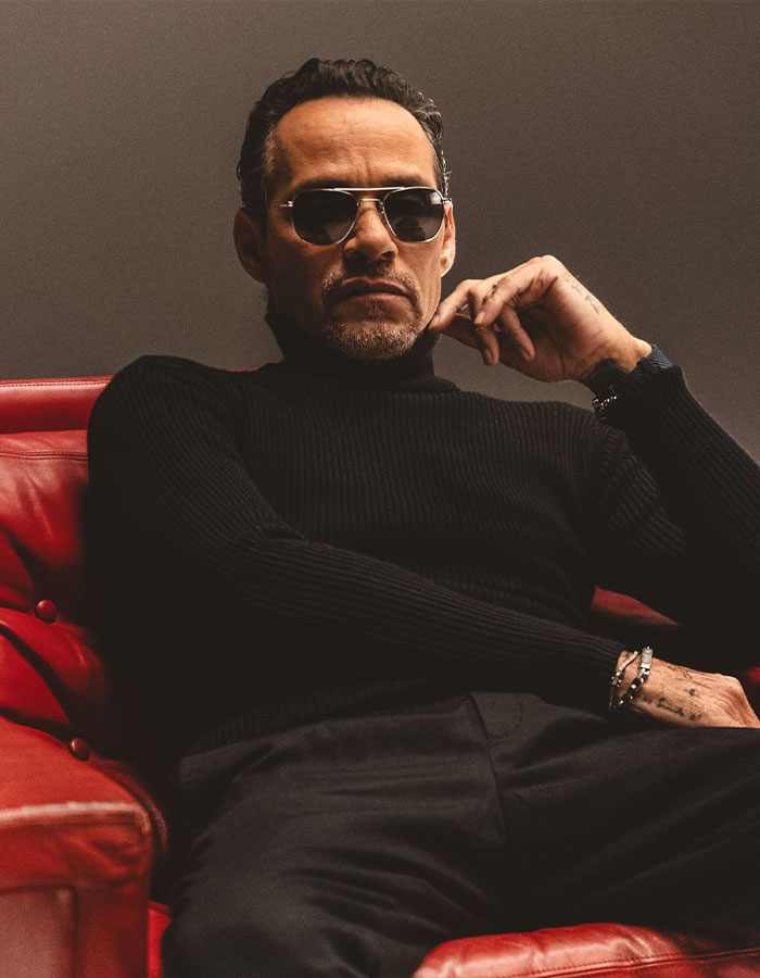 Marc Anthony sentado en sillón rojo con gafas oscuras y suéter negro, posando con expresión seria y confiada.