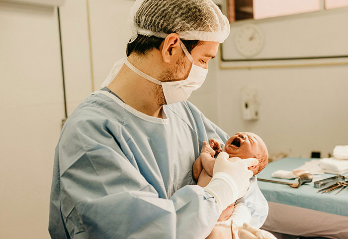 Médico en sala de parto sosteniendo bebé recién nacido mientras el caos por infidelidad comienza a surgir en el lugar.