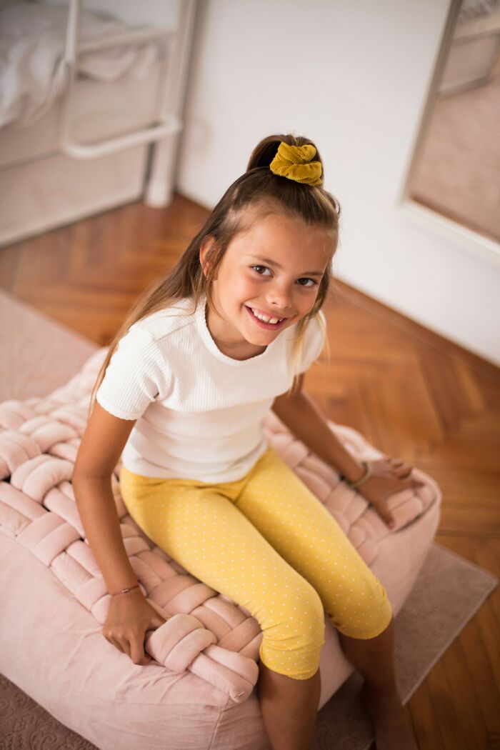 Niña sonriente con cabello recogido con scrunchie amarillo, sentada en puf rosa, mostrando expresión de karma real.