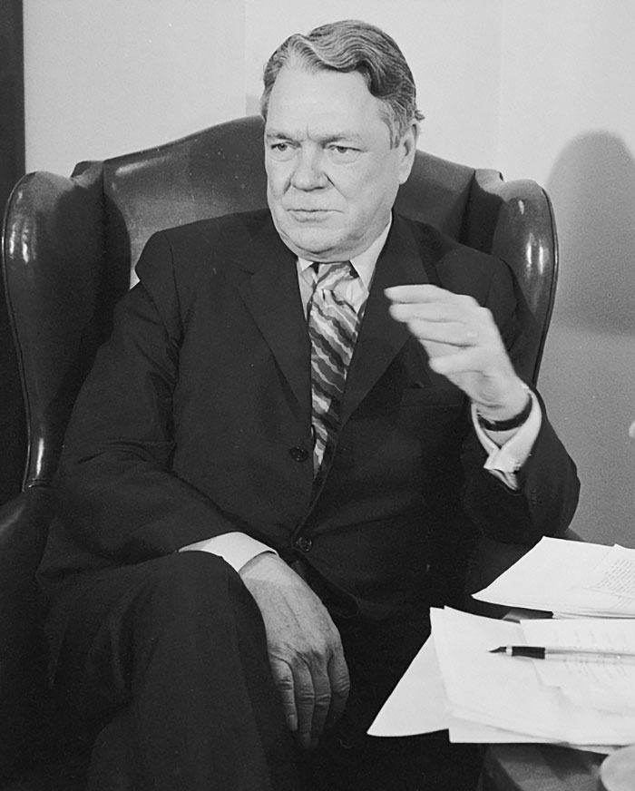 Figura pública sentada en sillón, vestida con traje y corbata, en una escena relacionada con figuras públicas que desaparecieron.