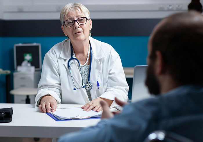 Mujer doctora con bata blanca y estetoscopio hablando con paciente masculino en consulta médica interior.