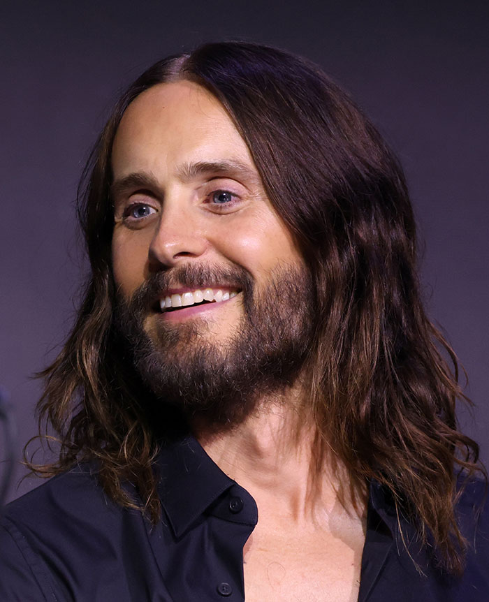 Hombre sonriente con cabello largo y barba, en entorno oscuro, mostrando una expresión amable y relajada. Hombre sonriente con cabello largo y barba, en entorno oscuro, mostrando una expresión amable y relajada.