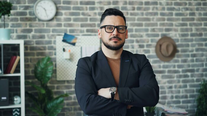 Hombre con gafas y chaqueta cruzando los brazos en oficina moderna mostrando expresión seria de karma real.