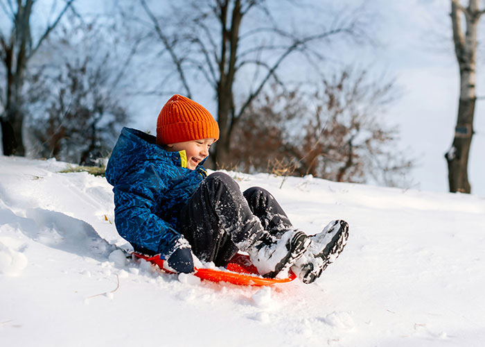 Niño feliz con gorro naranja y abrigo azul deslizándose en trineo sobre nieve, imagen relacionada con hallazgos autopsia médicos. Niño feliz con gorro naranja y abrigo azul deslizándose en trineo sobre nieve, imagen relacionada con hallazgos autopsia médicos.