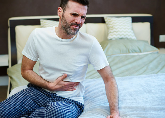 Hombre con dolor abdominal sentado en cama ilustrando síntomas relacionados con hallazgos sorprendentes en autopsias. Hombre con dolor abdominal sentado en cama ilustrando síntomas relacionados con hallazgos sorprendentes en autopsias.