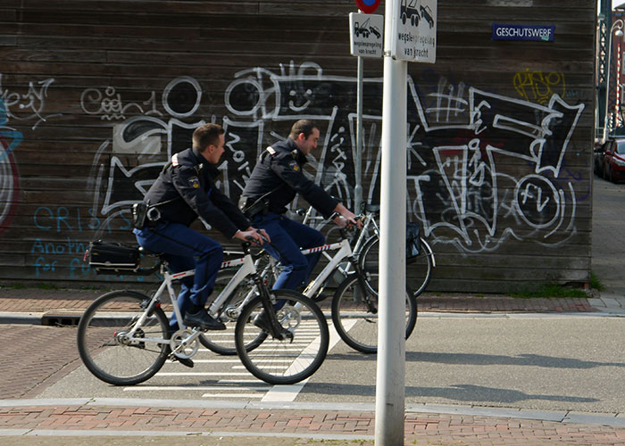 Dos policías montando bicicletas en una calle urbana con grafitis, mostrando un momento de karma instantáneo en acción.