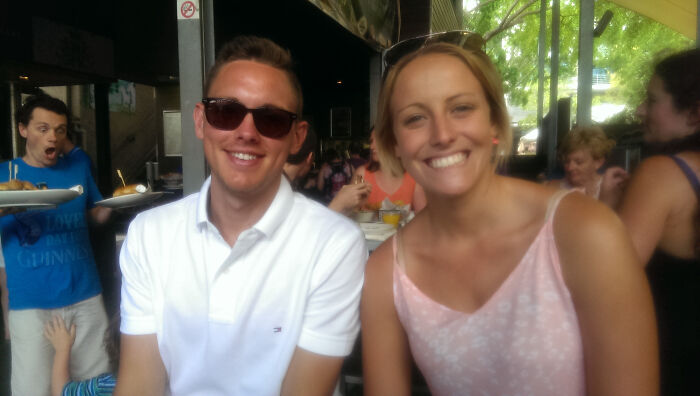 Pareja sonriente en restaurante al aire libre con cameo inesperado de hombre sorprendido detrás arruinando la foto divertida.