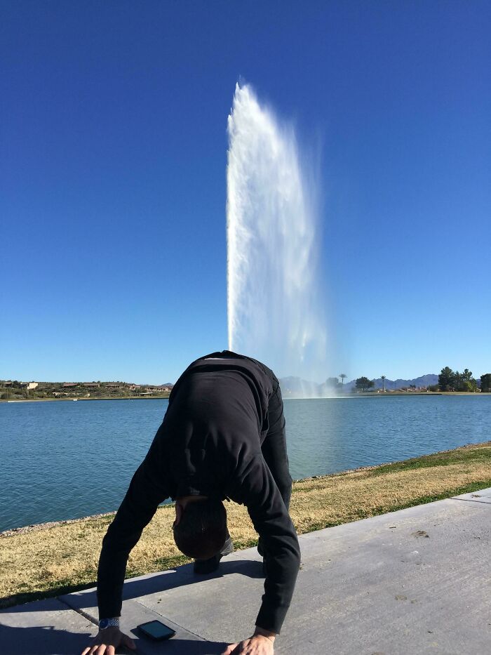 Hombre juguetón simulando que la fuente de agua brota de su cabeza en un día soleado cerca del lago.