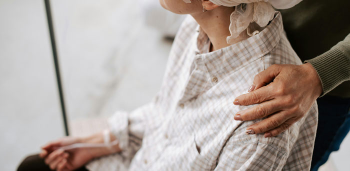 Persona mayor sentada con bata a cuadros mientras otra persona le ofrece apoyo tocando su hombro, mostrando drama familiar.