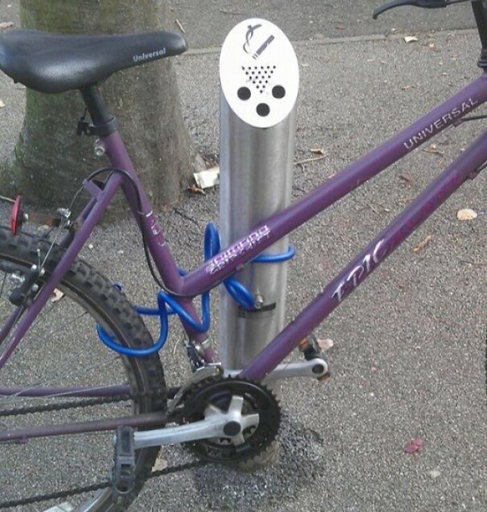 Bicicleta mal asegurada en poste para cigarrillos en espacio público mostrando falta de sentido común evidente.