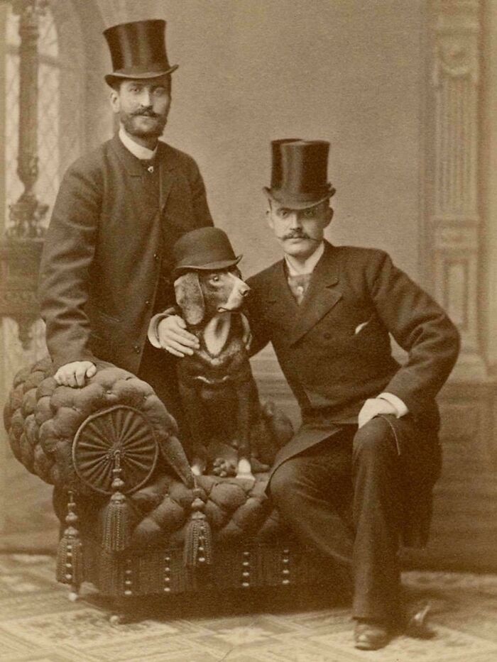Foto histórica rara y poderosa de dos hombres con sombreros de copa y un perro con sombrero en un sofá antiguo.