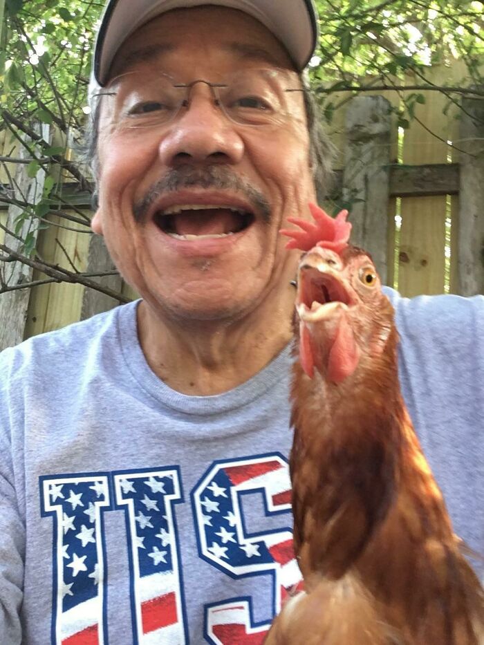 Hombre sonriente con gorra y camiseta de USA tomando selfie con una gallina en un entorno al aire libre.