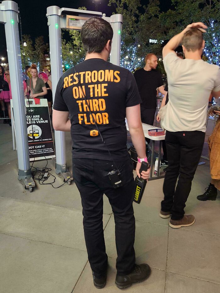 Hombre con camiseta indicando baños en tercer piso como solución ingeniosa a problemas cotidianos en evento nocturno.