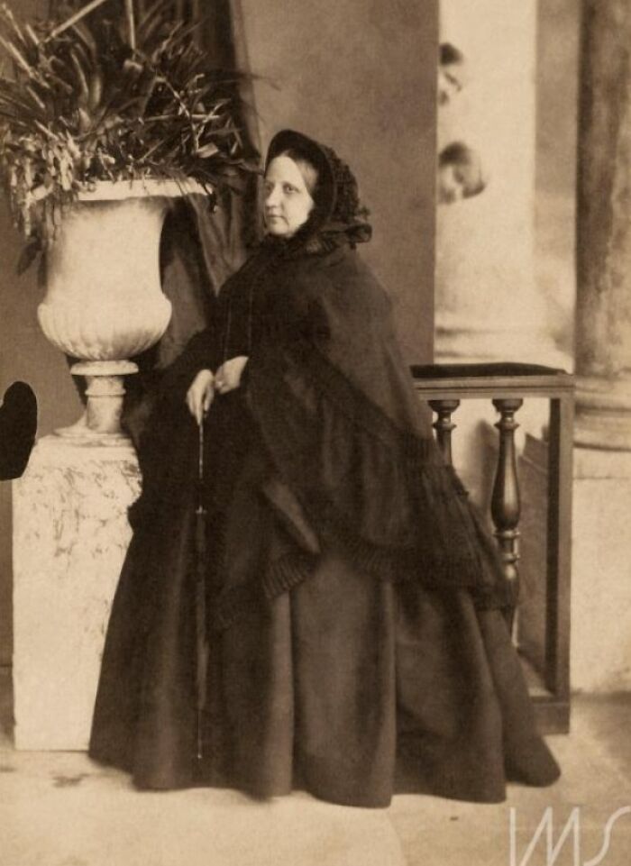 Mujer antigua posando vestida de negro mientras un cameo inesperado aparece detrás arruinando la foto divertida.