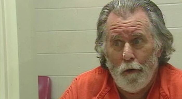 Hombre mayor con barba y cabello canoso, vistiendo uniforme naranja, en un entorno que sugiere detención o prisión.