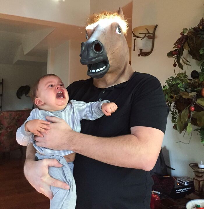 Hombre con máscara de caballo sostiene a un bebé que llora, mostrando momentos divertidos cuando hombres nunca crecieron.