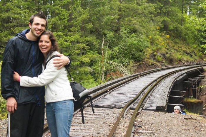 Pareja sonriente abrazándose cerca de vías de tren con cameo inesperado asomando detrás, fotos arruinadas divertidas.
