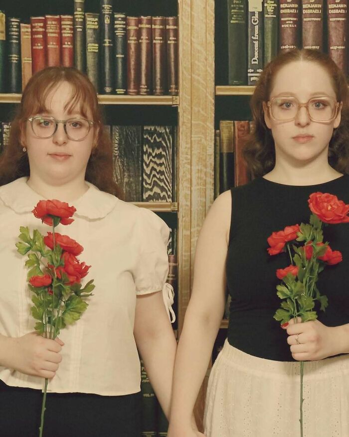 Dos mujeres con gafas sosteniendo flores rojas frente a una estantería de libros en una foto familiar fallida.