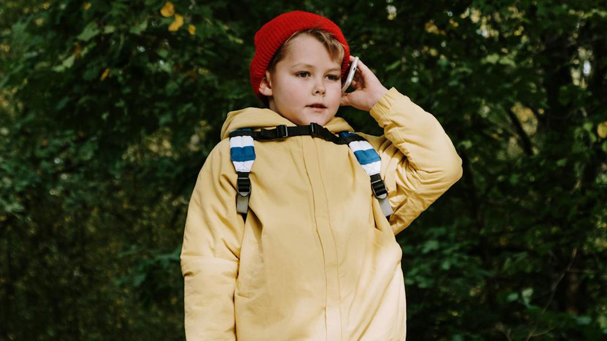 Niu00f1o con chaqueta amarilla y gorro rojo, usando un telu00e9fono mu00f3vil, representando momentos de padres con hijos difu00edciles.
