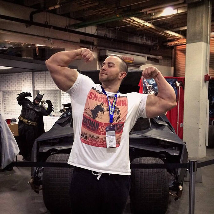 Hombre musculoso posando frente a un Batimóvil mientras Batman aparece inesperadamente en el fondo en cameo divertido.