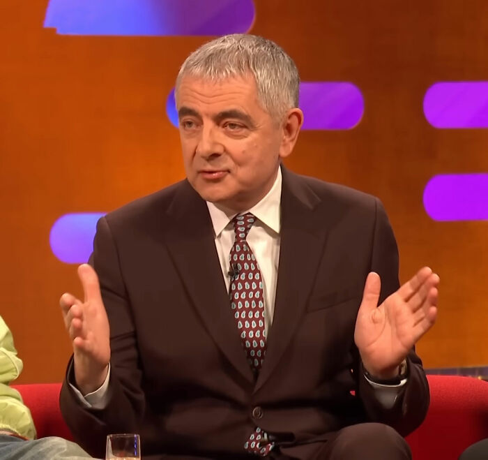 Hombre en traje oscuro gesticulando en un programa de entrevistas sobre mujeres corrigiendo a los chicos.