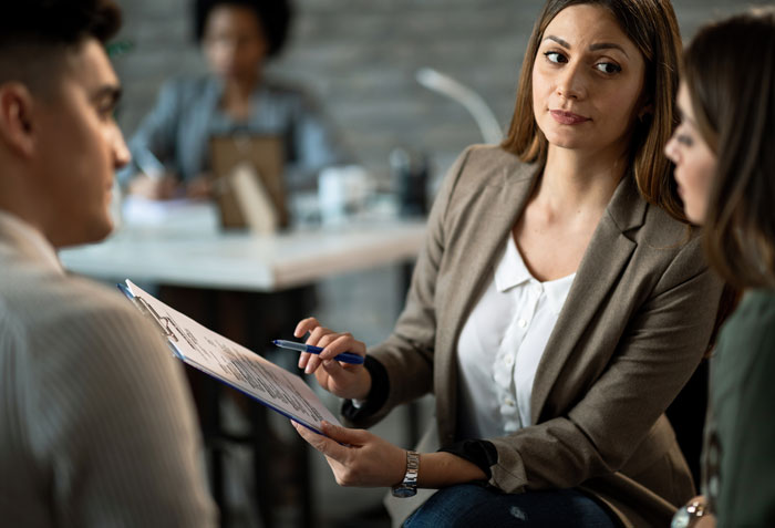 Mujer en oficina discutiendo un escándalo laboral mientras sostiene una carpeta con documentos frente a dos colegas atentos.