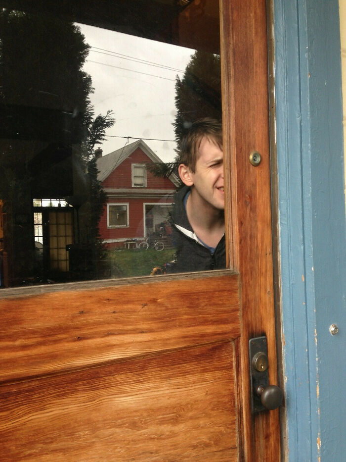 Persona mirando a través de una puerta de madera con reflexión de casa y árboles, imagen divertida de cosas inútiles encontradas.