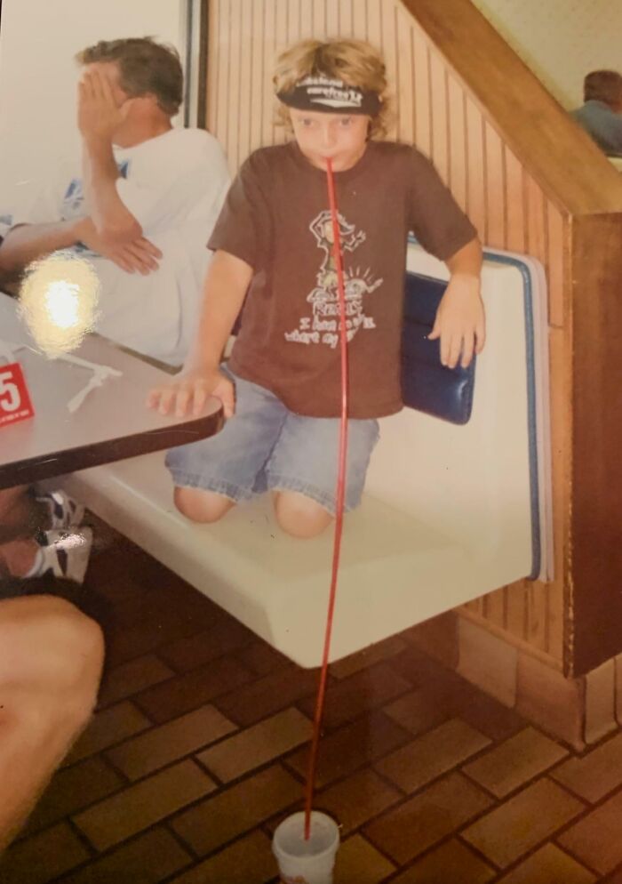 Niño con camiseta marrón usando una pajilla larga y hilarante en una foto infantil incómoda y divertida.