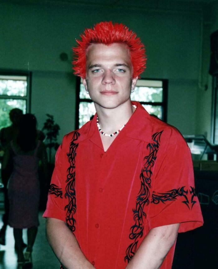 Joven con peinado punk de cabello rojo y camisa roja posando en una foto de infancia incómoda y divertida.