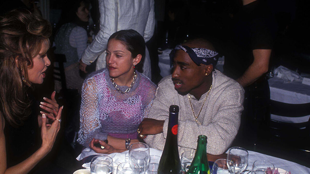 Foto impactante de cultura pop con personas famosas conversando en una cena social con ambiente oscuro y mesas con bebidas.